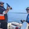 Student in glasses and hat smiles as he holds up a piece of equipment while on a boat on the water alongside a professor