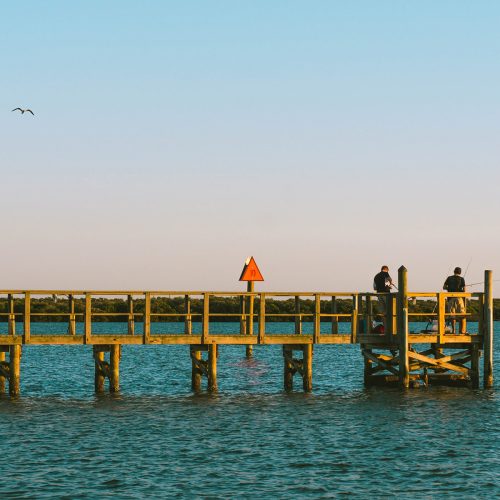 Two people fishing off the end of a long pier stretching into the bay