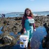 Woman in boots and waterproof pants standing in a tide pool while holding a big bucket