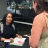 Woman seated at table signs a book while chatting with student