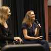 Two presenters sit at the edge of a stage while speaking into microphones