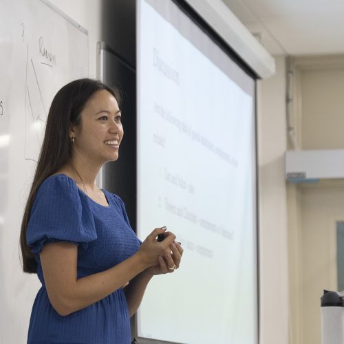 Professor in blue dress stands in front of dry erase board and screen at the front of a class