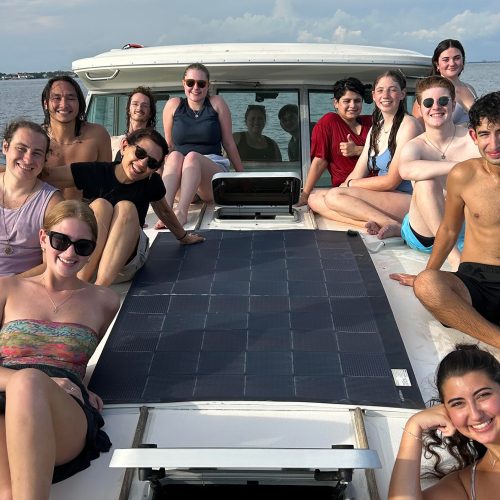 Twelve students sit on the deck of a boat next to solar panels