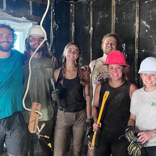 Group of seven in construction hats and gear stand in building with gutted walls