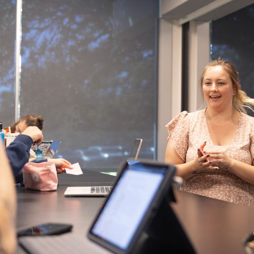 Professor stands in front of a desk where students listen and take notes on ipad