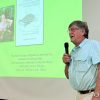 Man in glasses speaks into microphone in front of screen showing a photo of turtle researcher