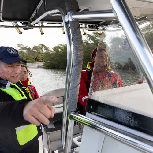 Man in brightly colored vest and hat on boat points to something afar as a student drives the boat