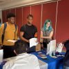 Four students stand around a table to talk to a pair of gentlemen